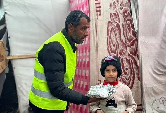A volunteer wearing a high-visibility vest hands a wrapped meal to a young child standing outside a tent.