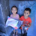 Two children stand in a modest indoor setting, holding a Giving Hands relief package.