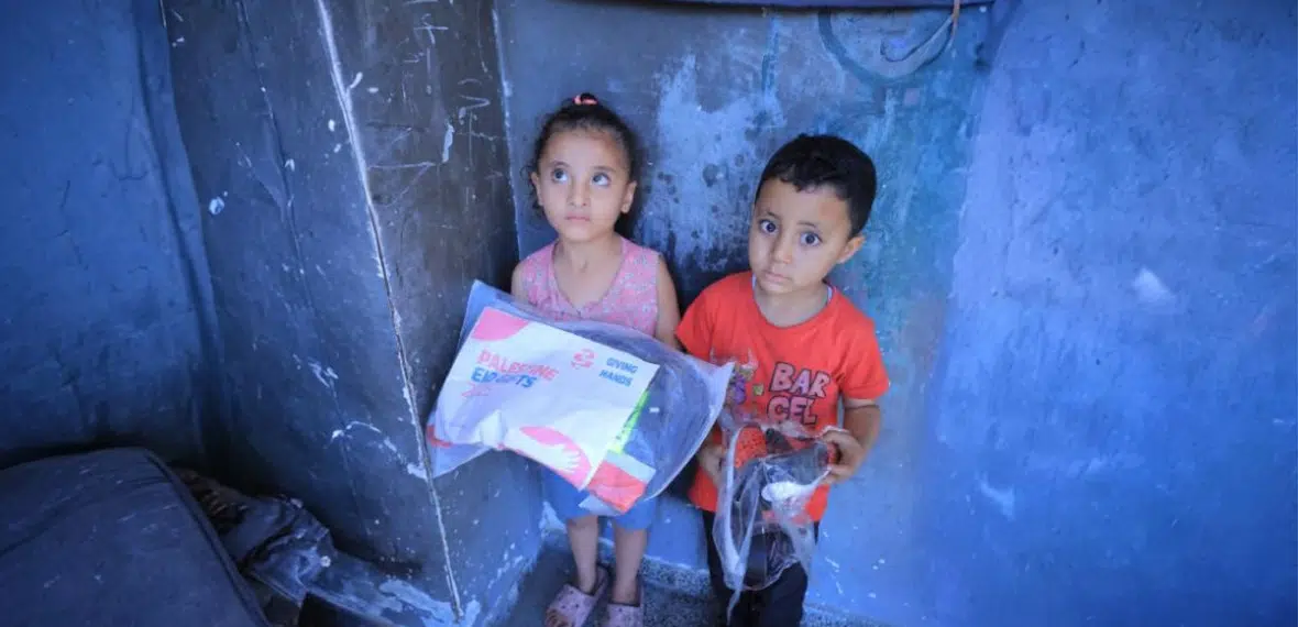 Two children stand in a modest indoor setting, holding a Giving Hands relief package.