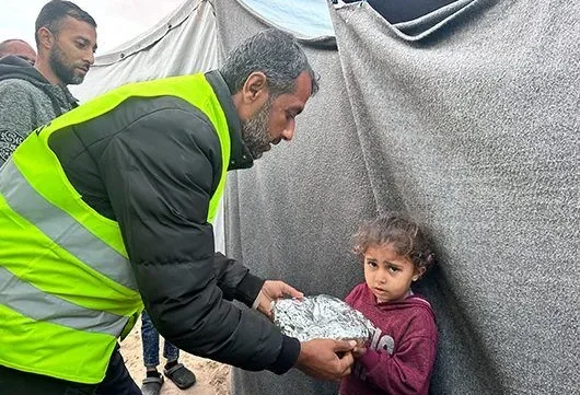 A Giving Hands volunteer in a yellow vest hands a wrapped meal to a young child standing beside a tent in a camp.