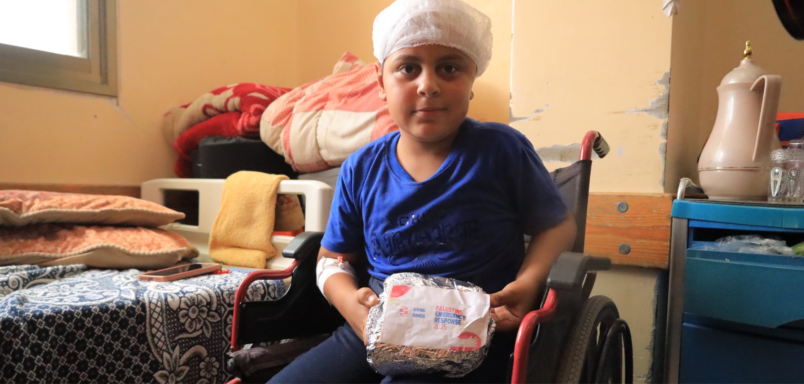 A person in a wheelchair holds an emergency food package labeled "Giving Hands Palestine Emergency Response 2025" in a modest room.