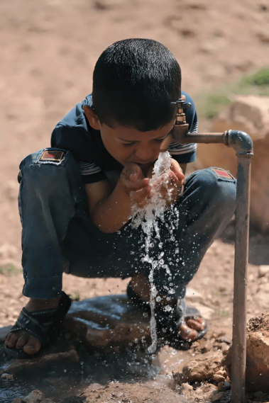 Child crouching and drinking water from a tap outdoors, with water splashing onto the ground