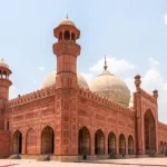 A large, historic mosque with red stone architecture and multiple domes, supported by Giving Hands.
