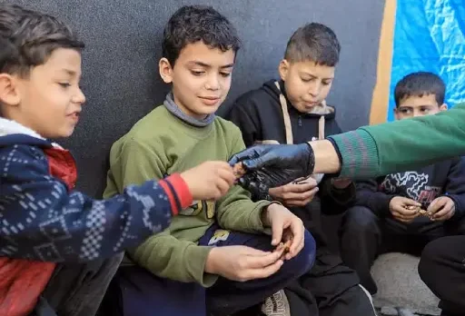 Several children sit together outdoors as a gloved hand distributes aid items to them.