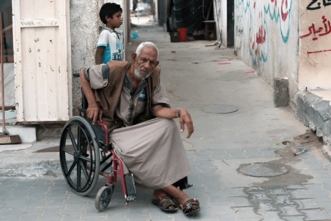 Man in a wheelchair sits on a paved alleyway while a child stands nearby, with graffiti on the walls in the background