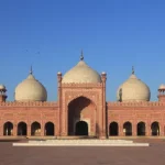 A grand mosque with three large domes and multiple minarets, built with red stone, supported by Giving Hands.