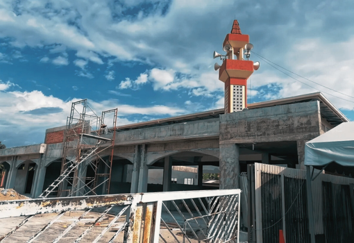 Mosque under construction with unfinished concrete walls, scaffolding, and a completed minaret beneath a blue sky