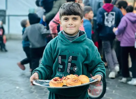 A child holds a tray of pastries at a Giving Hands food distribution event, with other children visible in the background.