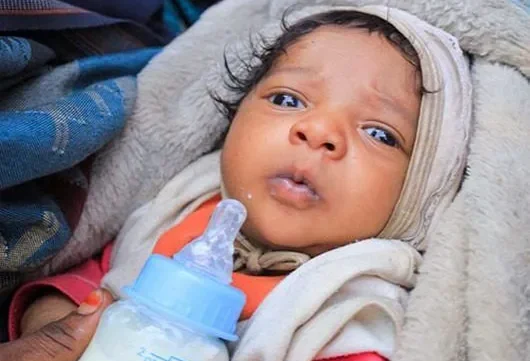 A caregiver holds a bottle to feed an infant wrapped in a blanket, providing nourishment and care.