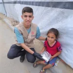 A Giving Hands volunteer sits beside a child outdoors, handing over a relief package.
