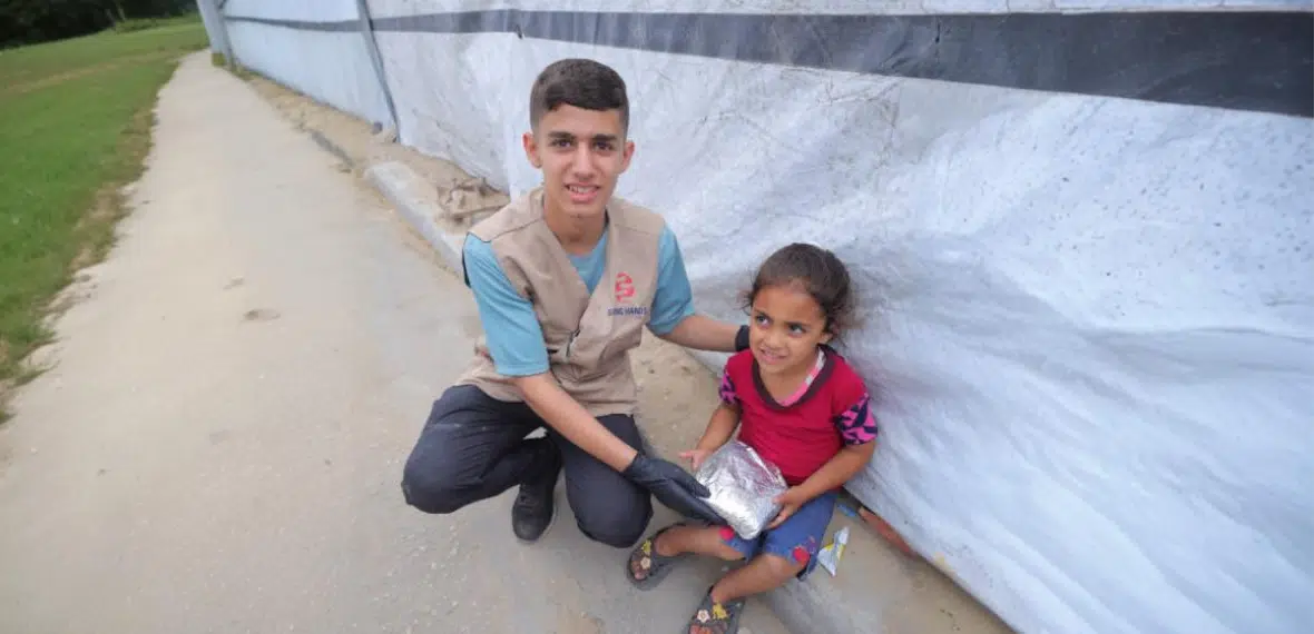 A Giving Hands volunteer sits beside a child outdoors, handing over a relief package.