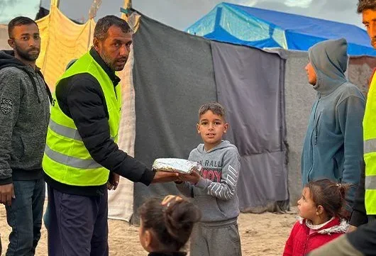 A Giving Hands volunteer in a yellow vest hands a wrapped meal to a child, while other children and adults stand nearby in a camp setting.