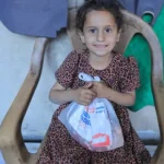 A young child sits on a chair holding a Giving Hands aid package in a modest indoor setting.