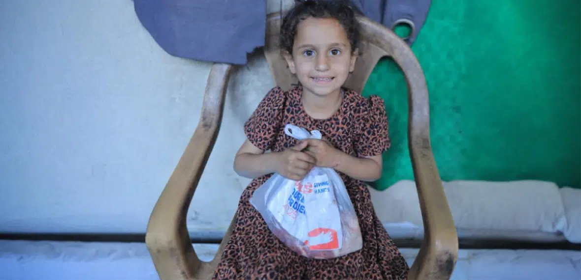 A young child sits on a chair holding a Giving Hands aid package in a modest indoor setting.