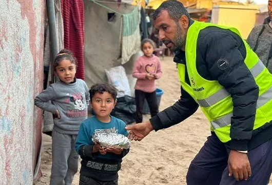 A Giving Hands volunteer in a yellow vest hands a wrapped meal to a child, with other children standing nearby in a camp setting.
