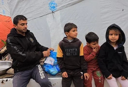 A volunteer distributes a blue emergency aid pack to a group of children standing outside a tent.