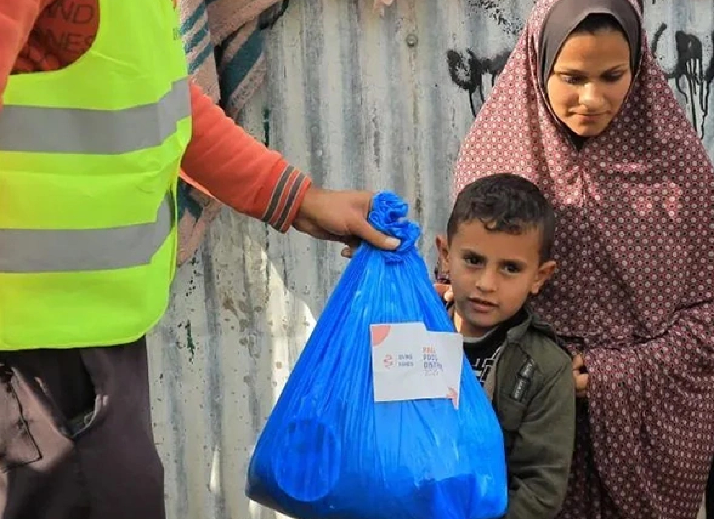 A volunteer from Giving Hands hands a blue bag of food supplies to a child and woman standing outdoors.