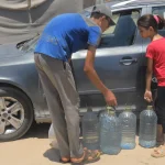 Two children fill large water containers next to a car during a Giving Hands emergency water aid distribution.