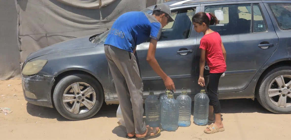 Two children fill large water containers next to a car during a Giving Hands emergency water aid distribution.