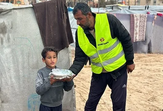 A Giving Hands volunteer in a yellow vest hands a wrapped meal to a child standing outside a tent in a camp setting.