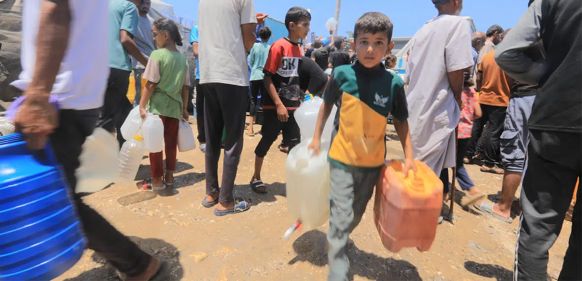 Children and adults carry water containers in a crowded outdoor setting during a Giving Hands humanitarian aid distribution.