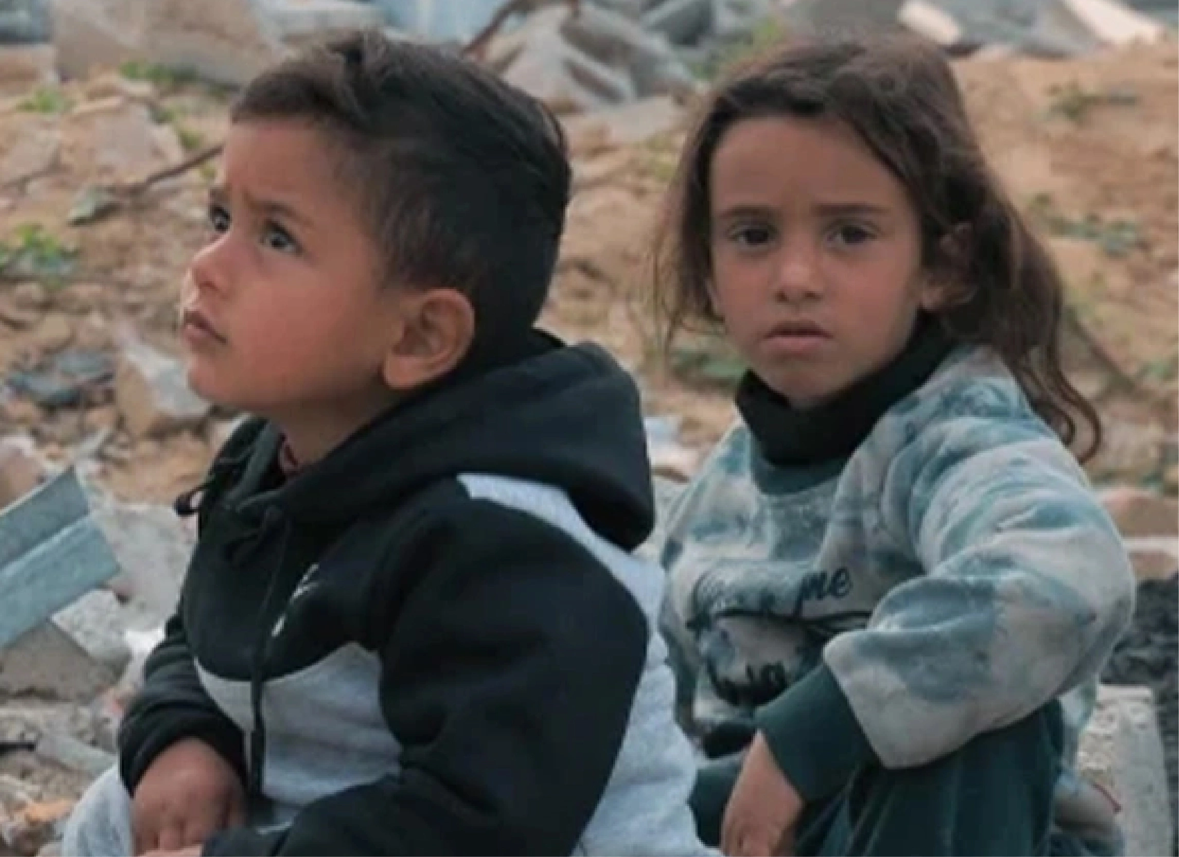 Two children sit among rubble and debris, supported by Giving Hands’ humanitarian relief efforts.
