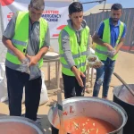 Volunteers in safety vests prepare large pots of food outdoors during a humanitarian aid event.
