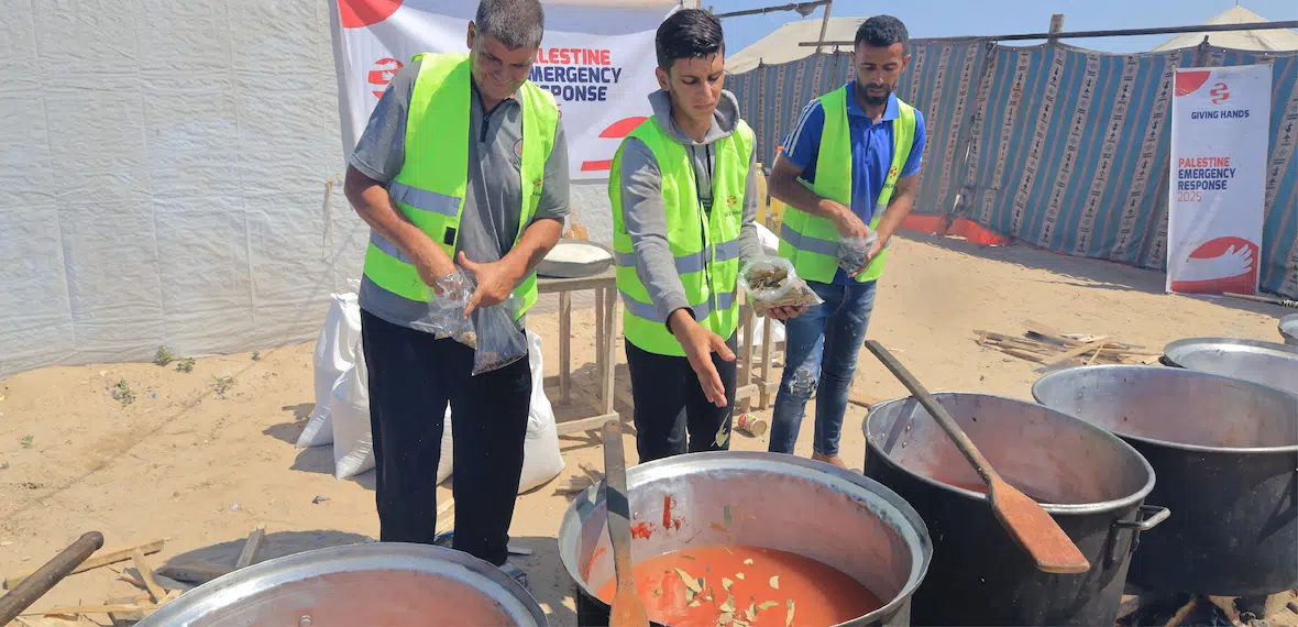 Volunteers in safety vests prepare large pots of food outdoors during a humanitarian aid event.
