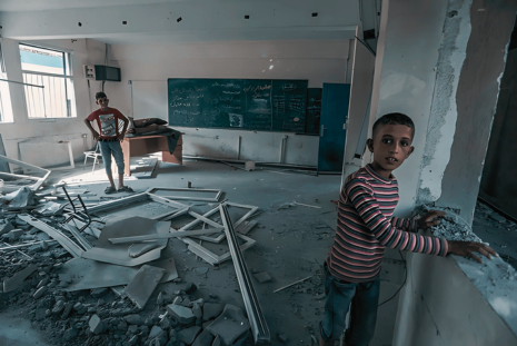 Children standing in a damaged classroom with debris, highlighting the need for rebuilding and educational support in Gaza