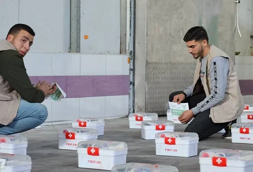 Two volunteers organize and distribute multiple first aid kits inside a building.
