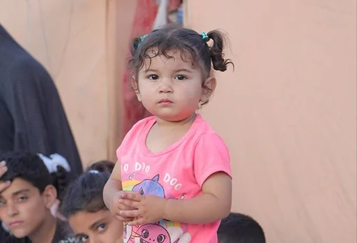 A young child in a pink shirt stands outside, with other children nearby.