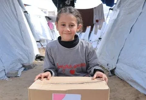 A young child holds a cardboard box in front of white tents at a refugee camp.
