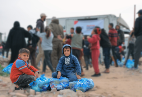 Two children sit on the ground with several blue food parcels in front of them, while a crowd gathers in the background during a relief distribution event.