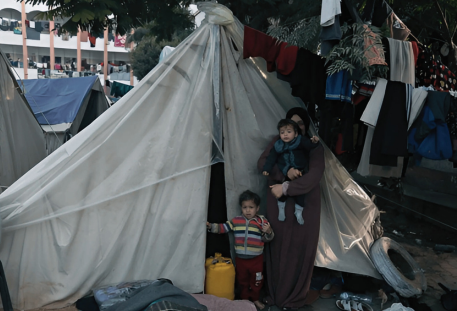 A woman stands with two young children outside a makeshift tent in a crowded camp, surrounded by belongings and supplies.