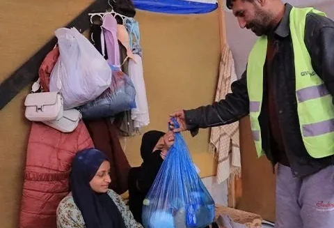 A person in a safety vest hands a blue bag of food to two people sitting inside a tent.