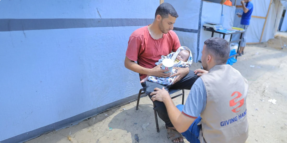 A givinghands aid worker provides baby milk formula to a father holding his infant outside a shelter in Gaza.