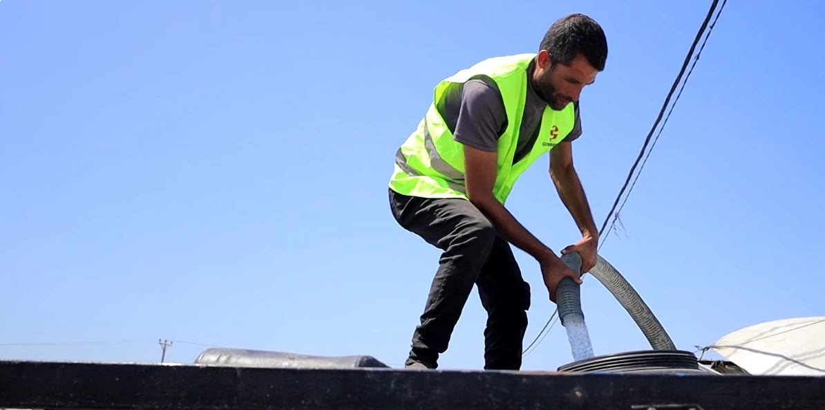 A relief worker in a safety vest fills a large water tank from a hose under a clear sky.