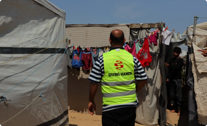 Giving Hands Canada aid worker providing humanitarian support in a Gaza refugee camp with tents and laundry hanging.