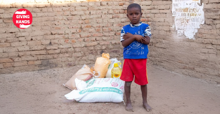 Child standing with food aid supplies provided by Giving Hands charity, including rice, flour, and cooking oil