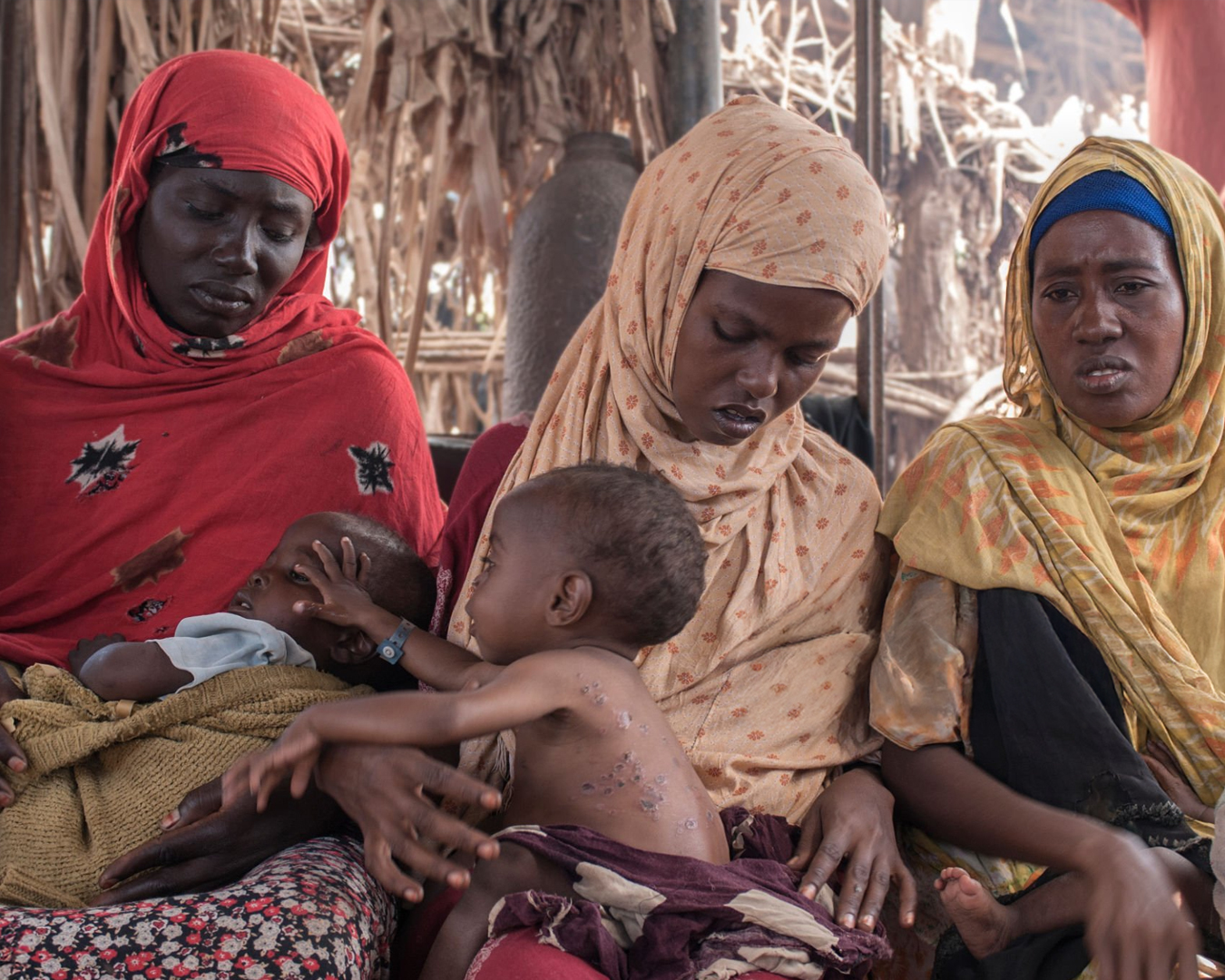 Sudanese women and children sitting together in a rural shelter, highlighting the need for emergency humanitarian aid