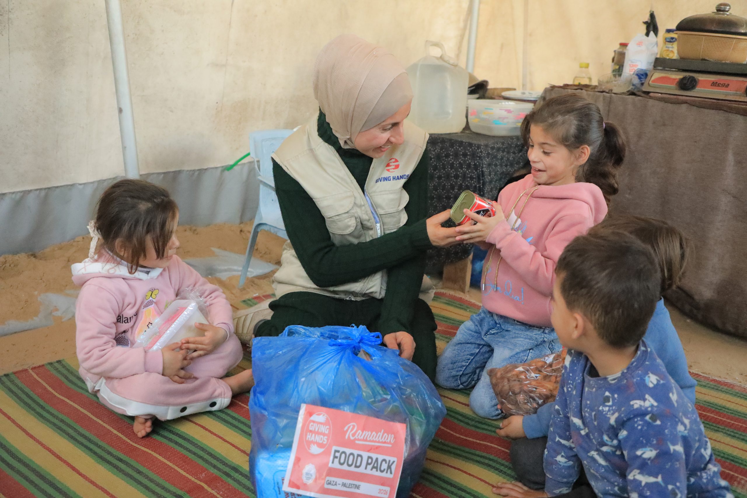 Giving Hands worker sitting with children inside a tent, sharing items from a Ramadan food pack