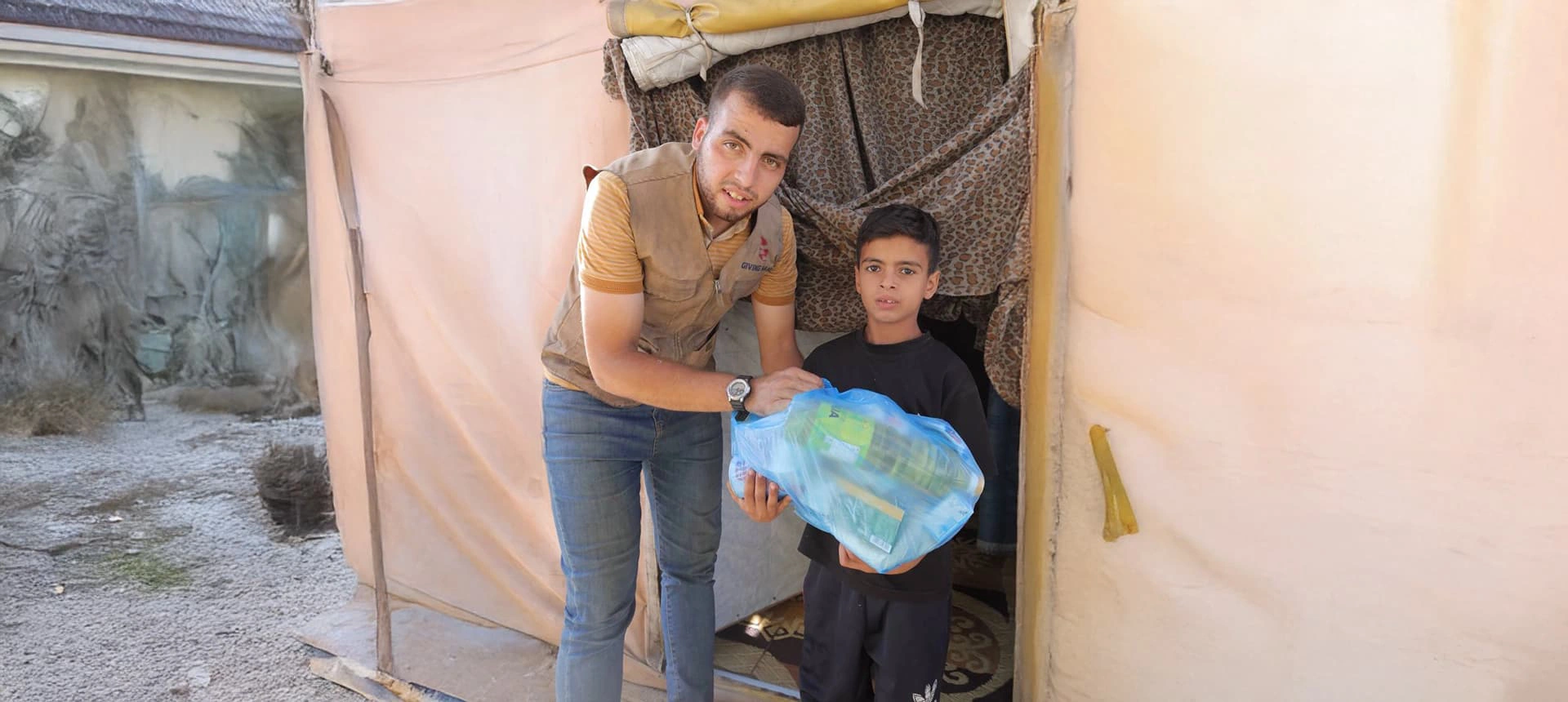 Giving Hands worker helping a child hold a blue food parcel at the entrance of a tent shelter