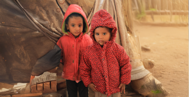 Two children in red hooded jackets standing together outside a tent in a dusty camp supported by Giving Hands