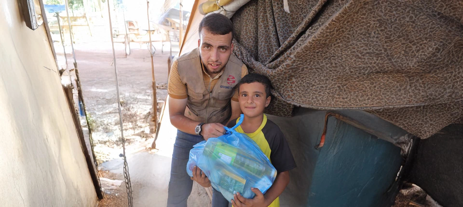 Giving Hands worker helping a child hold a blue food parcel at the entrance of a tent shelter