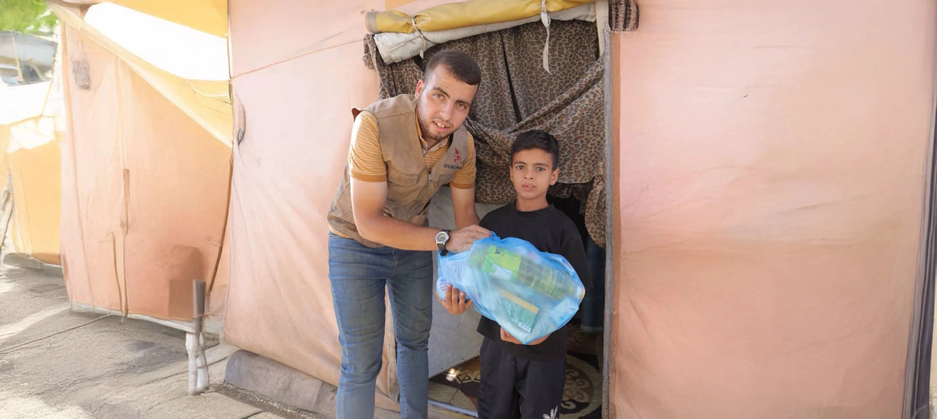 Giving Hands worker handing a blue food parcel to a child outside a tent shelter