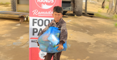 Child holding a large blue Ramadan food parcel in front of a Giving Hands banner