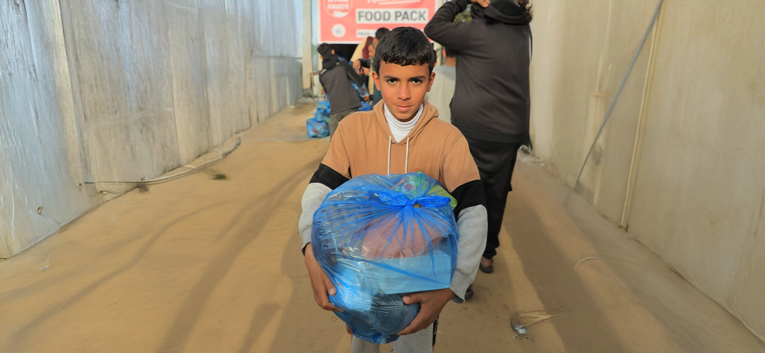 Child carrying a large blue food pack bag while walking through a narrow sandy corridor at a distribution point