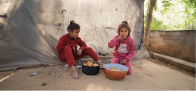 Two children sitting on sandy ground eating from plastic bowls in a makeshift shelter supported by Giving Hands