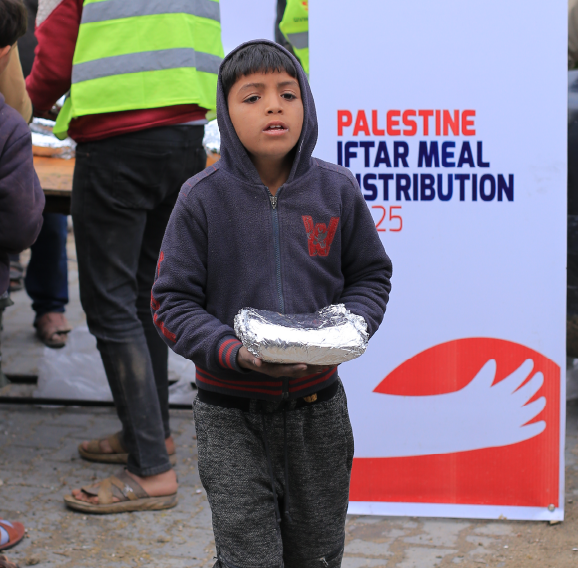 Child holding a foil-wrapped Iftar meal in front of a Palestine Iftar Meal Distribution 2025 banner