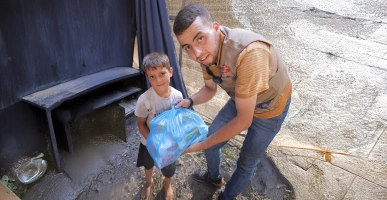 Giving Hands worker handing a blue food parcel to a barefoot child standing in a muddy area